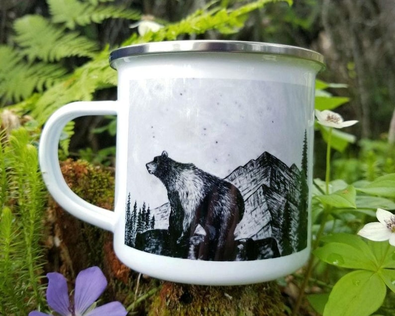 Puede incluir: Taza de camping de esmalte blanco con borde y asa plateados. La taza presenta una ilustraci&oacute;n en blanco y negro de un oso de pie frente a una cordillera y &aacute;rboles. La taza est&aacute; rodeada de follaje verde y flores.