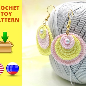 Op de afbeelding: Haak speelgoedpatroon om te downloaden. Een paar gehaakte oorbellen met een gelaagd ontwerp in geel en roze en een parel in het midden. De oorbellen hangen aan gouden haakjes. De oorbellen liggen op een bol grijs garen.
