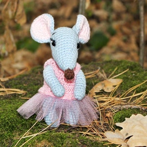 May include: A crocheted gray mouse wearing a pink dress and a tulle skirt. The mouse is sitting on a bed of moss and pine needles.