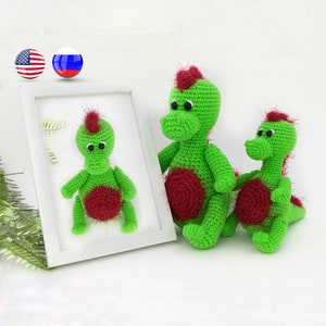 May include: Two green crocheted dinosaur toys with red accents. The larger dinosaur is sitting with its legs outstretched, while the smaller dinosaur is sitting with its tail curled around its legs.