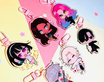 May include: Six colorful anime-style character keychains with pink metal rings. Each keychain features a different character with a unique design and expression. The keychains are displayed on a pink, yellow, and blue background.