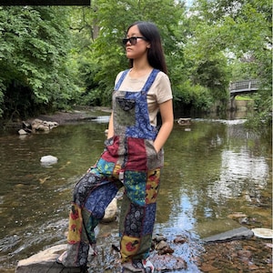 May include: A person wearing patchwork overalls with a beige t-shirt and sunglasses stands in a stream. The overalls feature a variety of colorful fabric patches in blue, red, green, and yellow. The image is set in a natural outdoor environment.