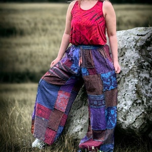 Może przedstawiać: Kobieta w czerwonej koszulce bez rękawów i spodniach z patchworku stoi na polu z dużym kamieniem za sobą. Spodnie są wykonane z różnych tkanin i kolorów, w tym niebieskiego, fioletowego i brązowego.