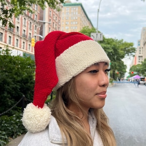 K&ouml;nnte beinhalten: Eine rot-wei&szlig;e gestrickte Weihnachtsm&uuml;tze mit einem wei&szlig;en Bommel oben. Die M&uuml;tze wird von einer Person mit langen blonden Haaren getragen.