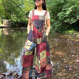 May include: A colourful patchwork jumpsuit featuring red, green, and blue panels. The wide-leg overalls have a patchwork design with various patterns. The model is wearing a white t-shirt and black sandals. The background is a natural outdoor setting.