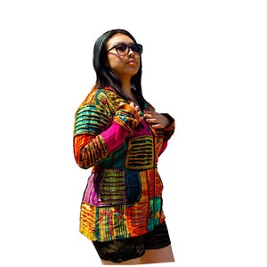 Może przedstawiać: Kobieta w kolorowej kurtce w stylu patchwork z zamkiem błyskawicznym. Kurtka ma różne kolory i wzory, w tym czerwony, żółty, zielony, niebieski i fioletowy. Kobieta ma na sobie czarne spodenki i okulary przeciwsłoneczne.