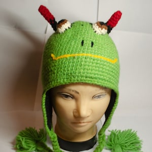 May include: Green crocheted hat with frog features, including eyes, a mouth, and horns. The hat has ear flaps and a pom-pom on each flap.