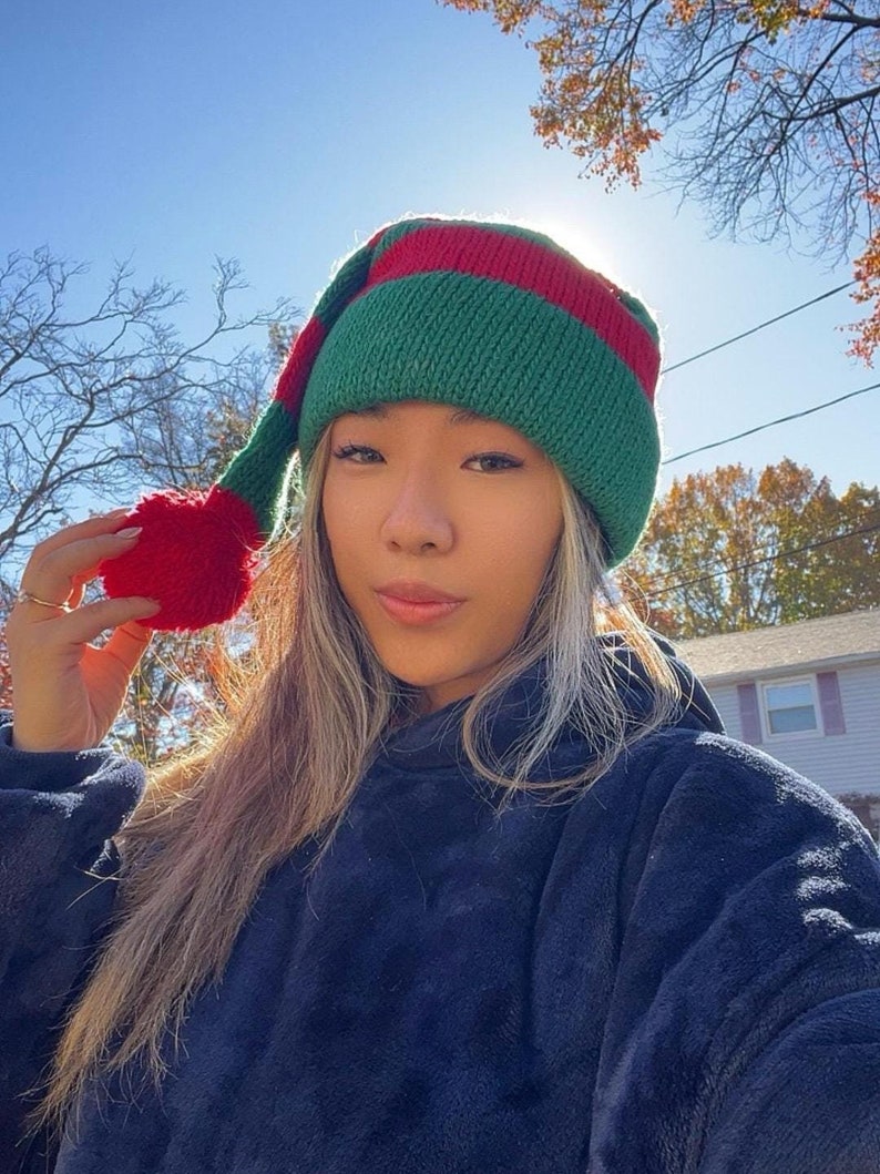 K&ouml;nnte beinhalten: Eine Frau tr&auml;gt eine gr&uuml;n-rot gestreifte Elfenm&uuml;tze mit einem roten Bommel oben. Die M&uuml;tze hat eine lange, spitze Spitze. Sie tr&auml;gt einen dunkelblauen Fleece-Hoodie.