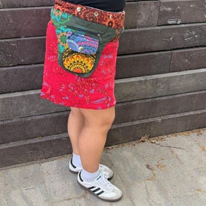 Puede incluir: Una falda envolvente roja con un estampado floral colorido y una ri&ntilde;onera a juego. La ri&ntilde;onera tiene m&uacute;ltiples bolsillos y un dise&ntilde;o decorativo. La falda se combina con zapatillas blancas con rayas negras.
