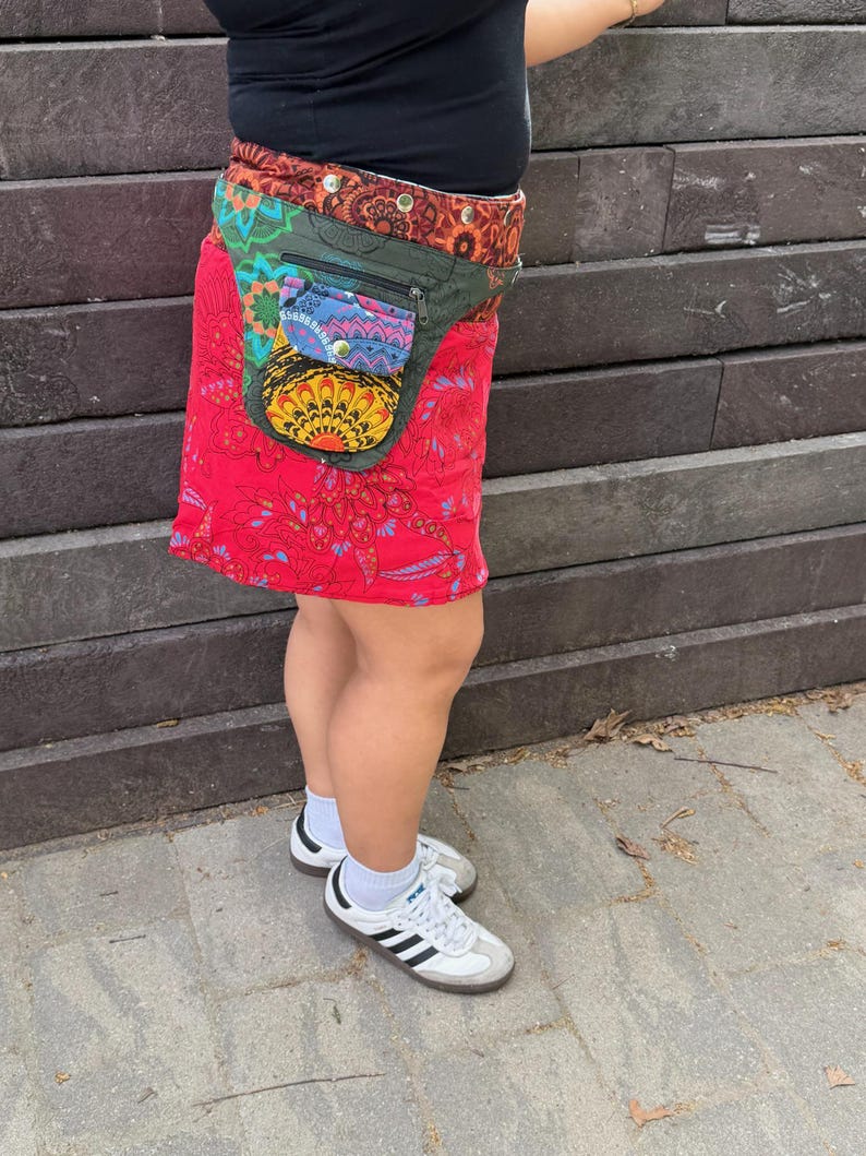 Puede incluir: Una falda envolvente roja con un estampado floral colorido y una ri&ntilde;onera a juego. La ri&ntilde;onera tiene m&uacute;ltiples bolsillos y un dise&ntilde;o decorativo. La falda se combina con zapatillas blancas con rayas negras.