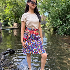 Peut inclure: Une femme porte un t-shirt beige et une jupe portefeuille colorée dans un ruisseau peu profond. La jupe présente un motif vibrant de cercles bleus, rouges et oranges. Elle porte des lunettes de soleil et des sandales, avec un fond vert naturel.