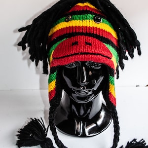 May include: A black knit hat with red, yellow, and green stripes. The hat has a face with black eyes and a black nose. The hat has long black dreadlocks and two long black tassels.