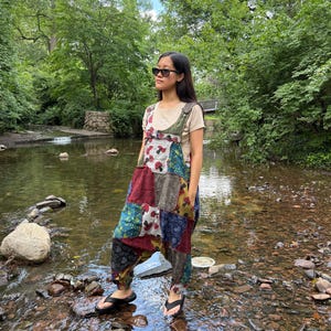 May include: A colourful patchwork jumpsuit with red, blue, and green patterns. The jumpsuit is worn with a light-coloured t-shirt and black sandals. The person is standing in a stream with a natural background of trees and water.