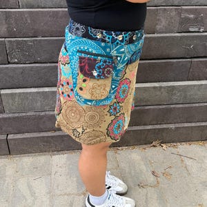 Puede incluir: Una falda envolvente colorida con un estampado floral y paisley en tonos azules, beige, rosa y marr&oacute;n. La falda tiene un bolsillo con botones y una cremallera. La persona lleva zapatillas blancas con rayas negras.