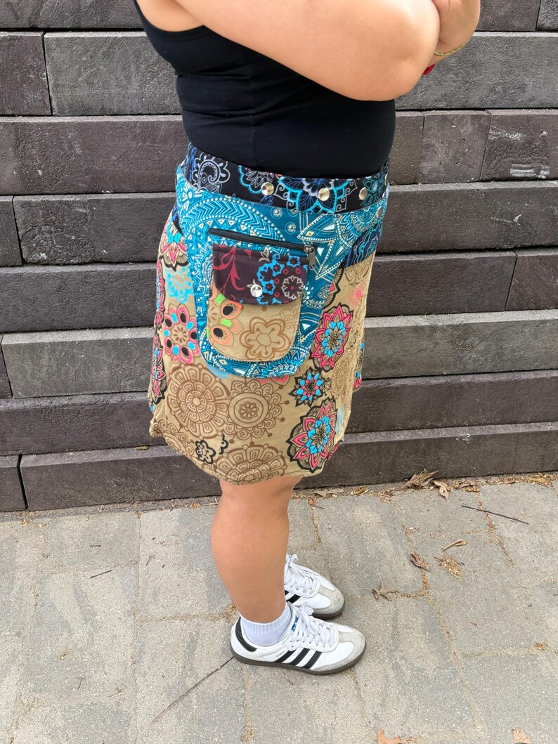 Puede incluir: Una falda envolvente colorida con un estampado floral y paisley en tonos azules, beige, rosa y marr&oacute;n. La falda tiene un bolsillo con botones y una cremallera. La persona lleva zapatillas blancas con rayas negras.