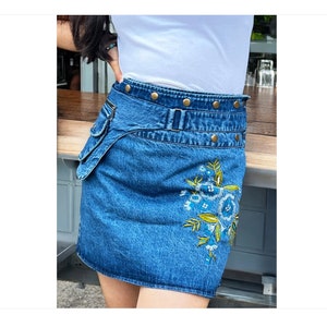 Puede incluir: Una falda vaquera azul con un diseño de bordado floral. La falda tiene un cinturón doble con tachuelas doradas y un bolsillo de utilidad en el lateral.