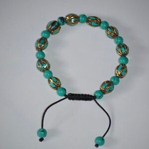 May include: A turquoise and gold beaded bracelet with a black cord closure. The beads are round and have a decorative design.
