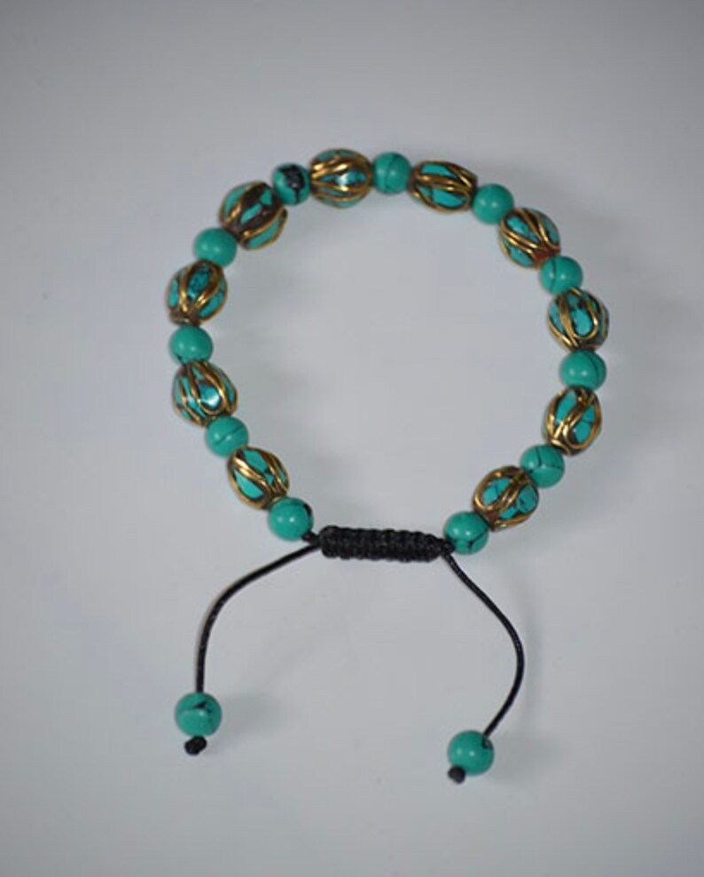 May include: A turquoise and gold beaded bracelet with a black cord closure. The beads are round and have a decorative design.