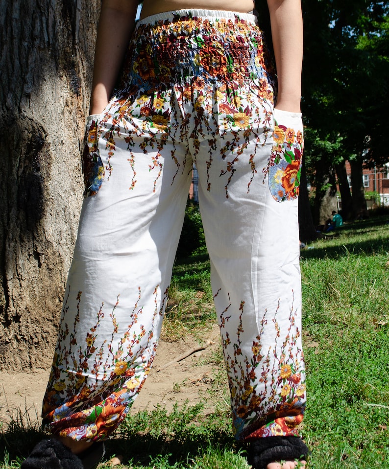 May include: White pants with a floral print featuring brown, orange, and yellow flowers. The pants have a wide leg and a drawstring waist.
