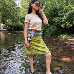Peut inclure: Une femme porte un t-shirt beige, une jupe à motifs et des lunettes de soleil dans un ruisseau. La jupe est principalement verte avec un motif circulaire répété. Une ceinture colorée accentue la taille. Des sandales noires complètent la tenue.