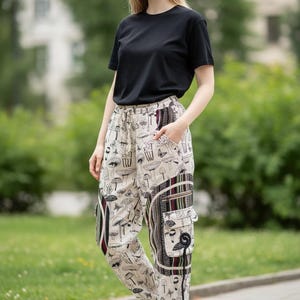 May include: A person wearing a black t-shirt and beige pants with a mushroom print. The pants have cargo pockets and a drawstring waist. White sneakers complete the outfit. The background is a blurred outdoor scene.
