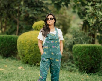 May include: A pair of teal denim overalls featuring a tie-dye pattern with purple and yellow floral accents, worn over a plain white short-sleeved t-shirt. The overalls include a front bib pocket, side buttons, and full-length pant legs.