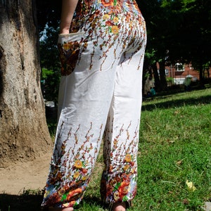 May include: White pants with a floral print featuring brown, orange, and yellow flowers. The pants have a wide leg and a loose fit.