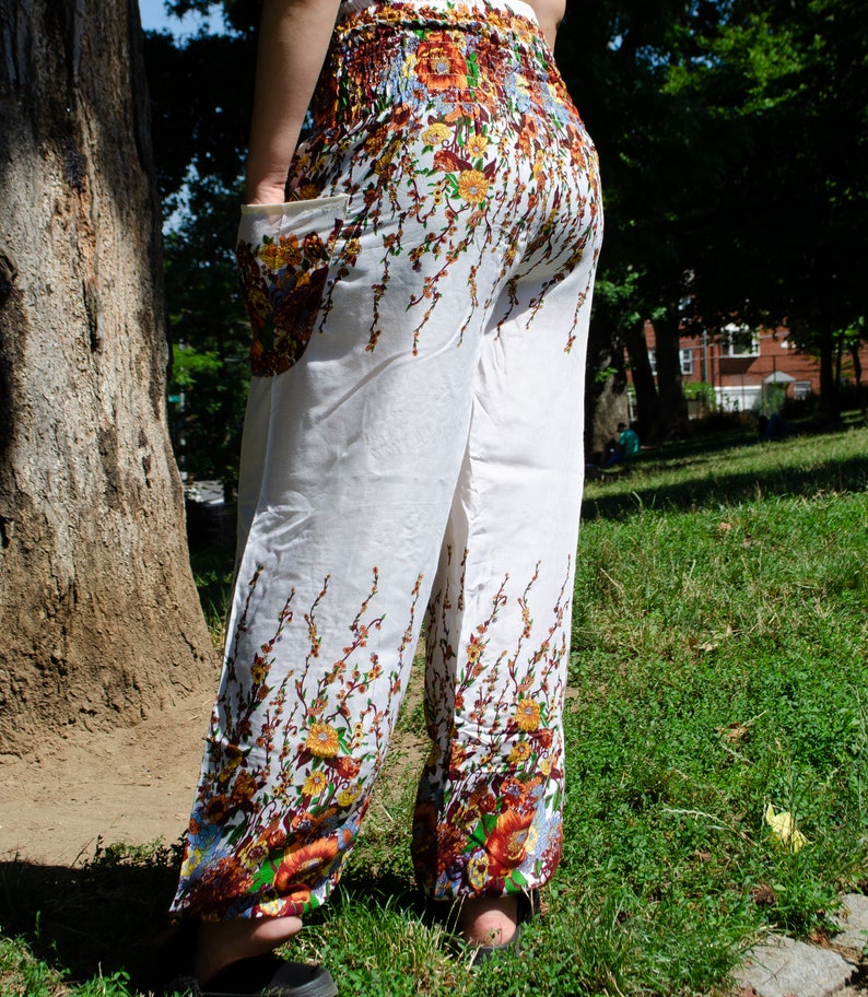 May include: White pants with a floral print featuring brown, orange, and yellow flowers. The pants have a wide leg and a loose fit.