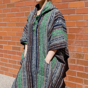 May include: A person wearing a green, brown, and black striped hooded poncho with fringe. The poncho has a pocket on each side. The person is wearing sunglasses and has their hair pulled back in a bun.
