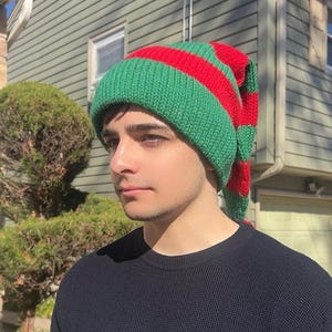 K&ouml;nnte beinhalten: Eine gr&uuml;n-rot gestreifte Strickm&uuml;tze mit einer langen, spitzen Spitze. Die M&uuml;tze ist aus einem weichen, warmen Material gefertigt und perfekt, um Kopf und Ohren im Winter warm zu halten.