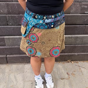 Puede incluir: Una mujer con una camiseta sin mangas negra, una falda estampada y un cintur&oacute;n con bolsillo. La falda es de color beige con grandes dise&ntilde;os florales en rosa y azul. El cintur&oacute;n es azul y negro con detalles plateados. Zapatillas blancas completan el atuendo.
