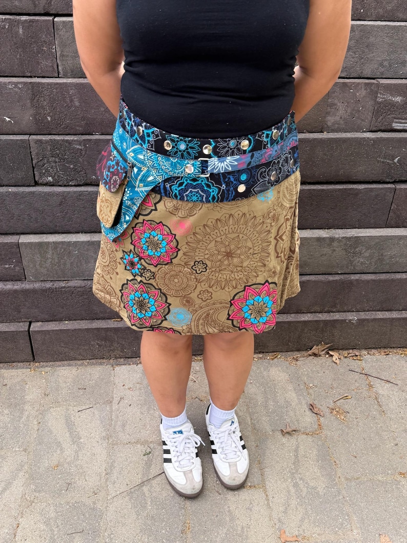 Puede incluir: Una mujer con una camiseta sin mangas negra, una falda estampada y un cintur&oacute;n con bolsillo. La falda es de color beige con grandes dise&ntilde;os florales en rosa y azul. El cintur&oacute;n es azul y negro con detalles plateados. Zapatillas blancas completan el atuendo.