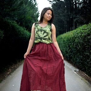 Puede incluir: Una mujer con un top de tirantes de color verde y blanco con estampado tie-dye y una falda larga de color burdeos. Está de pie en un camino pavimentado bordeado de arbustos verdes.