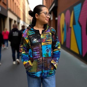 May include: A colorful patchwork jacket with a hood and zipper. The jacket features a vibrant mix of blues, greens, yellows, and purples in a tie-dye style. The person is wearing blue jeans.