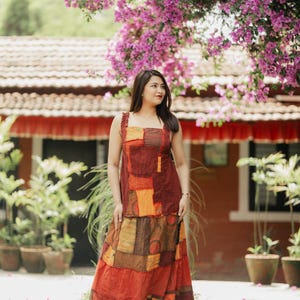 May include: A sleeveless dress featuring a patchwork design with various rectangular fabric panels in shades of deep red, burnt orange, and mustard yellow. The bodice has wide straps and a square neckline, while the skirt is long and flowing.