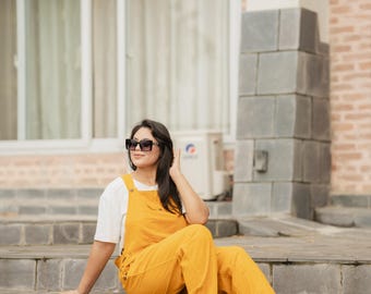 May include: A pair of mustard yellow denim overalls worn over a plain white short-sleeved t-shirt, paired with white sneakers and dark-framed sunglasses. The overalls feature a front bib pocket, side button closures, and wide-leg trousers.