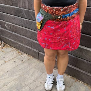 Puede incluir: Una falda envolvente roja con estampado de cachemir, combinada con un cintur&oacute;n multicolor con un peque&ntilde;o bolsillo. El cintur&oacute;n tiene un bolsillo verde con un dise&ntilde;o colorido. El conjunto se completa con zapatillas blancas y una camiseta negra.