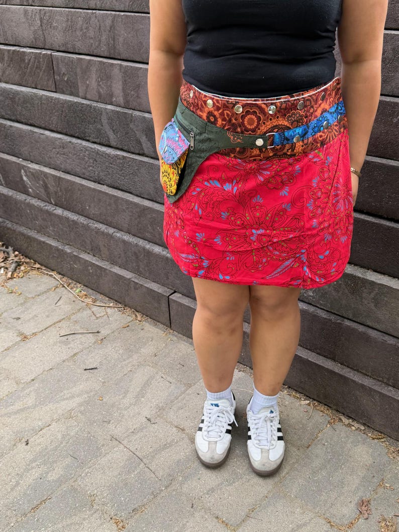 Puede incluir: Una falda envolvente roja con estampado de cachemir, combinada con un cintur&oacute;n multicolor con un peque&ntilde;o bolsillo. El cintur&oacute;n tiene un bolsillo verde con un dise&ntilde;o colorido. El conjunto se completa con zapatillas blancas y una camiseta negra.