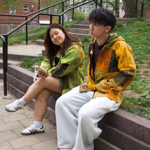 May include: Two people are seated outdoors, wearing colorful patchwork jackets. One jacket is green and black, the other is yellow and orange. Both are wearing white pants and sneakers. The setting is a park with a brick walkway and a grassy area.
