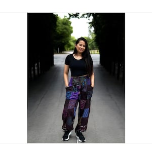 May include: A woman wearing a black crop top and colorful patchwork pants stands in a tree-lined path. She is smiling and looking at the camera.