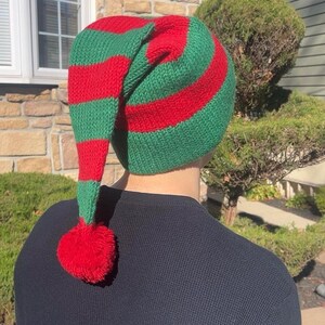 K&ouml;nnte beinhalten: Eine gr&uuml;n-rot gestreifte gestrickte Elfenm&uuml;tze mit einem roten Bommel am Ende der langen, spitzen Spitze.