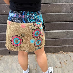Puede incluir: Falda envolvente con base beige, con un estampado floral y paisley en tonos rosa, azul y turquesa. La cintura es una mezcla de patrones azules y negros. La falda se lleva con una camiseta sin mangas negra y zapatillas blancas.