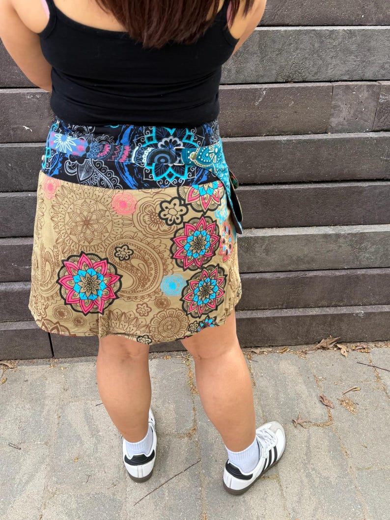 Puede incluir: Falda envolvente con base beige, con un estampado floral y paisley en tonos rosa, azul y turquesa. La cintura es una mezcla de patrones azules y negros. La falda se lleva con una camiseta sin mangas negra y zapatillas blancas.