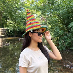 May include: A colorful, striped crochet hat with a pointed top and wide brim. The hat features bands of red, yellow, green, and orange. The hat is worn outdoors, with a natural background of trees and water.