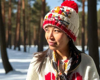 Puede incluir: Un gorro de invierno tejido con un borde rojo y un gran pompón rojo. El gorro es de color crema con un estampado floral en rojo, amarillo y negro. La persona lleva un suéter color crema con un diseño de corazón rojo.