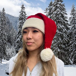 K&ouml;nnte beinhalten: Eine rot-wei&szlig;e gestrickte Weihnachtsm&uuml;tze mit einem wei&szlig;en Bommel oben. Die M&uuml;tze wird von einer Person mit langen blonden Haaren getragen, die vor einem verschneiten Waldhintergrund steht.