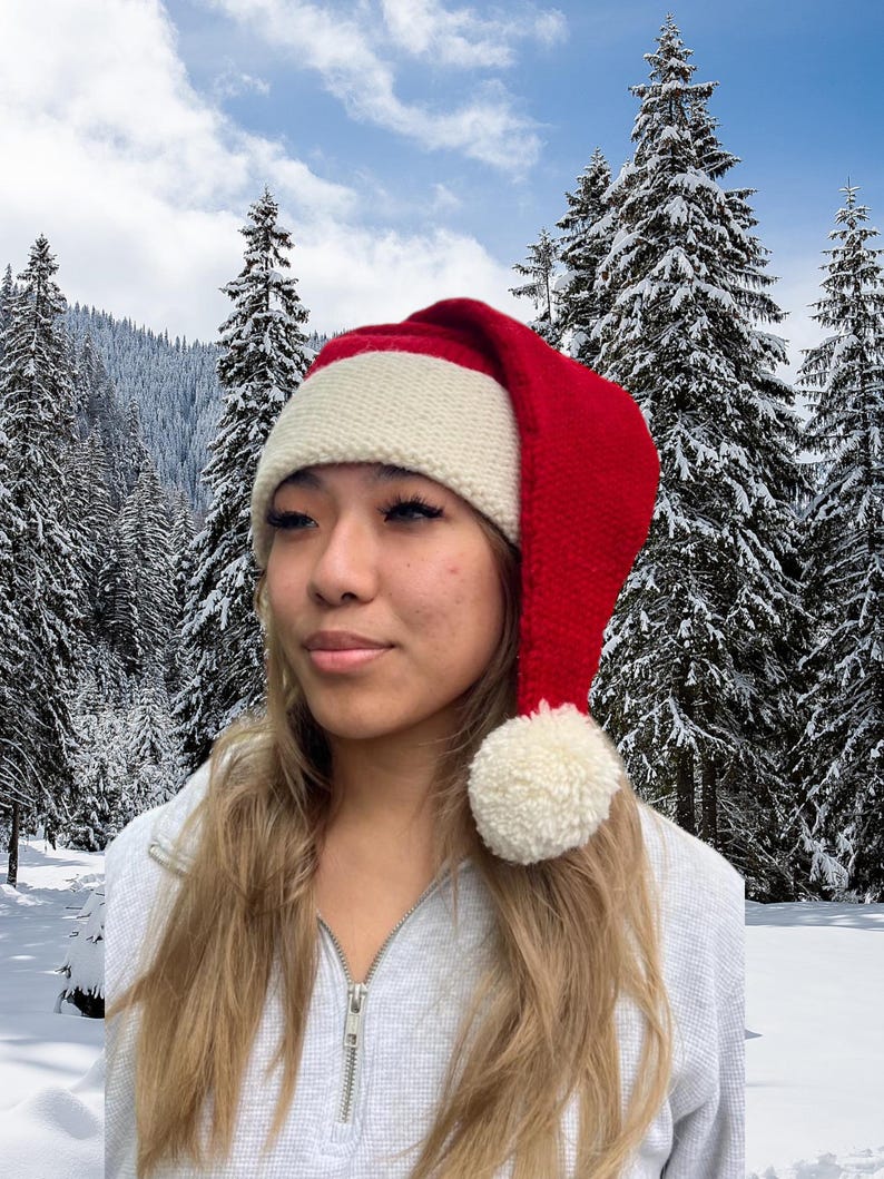 K&ouml;nnte beinhalten: Eine rot-wei&szlig;e gestrickte Weihnachtsm&uuml;tze mit einem wei&szlig;en Bommel oben. Die M&uuml;tze wird von einer Person mit langen blonden Haaren getragen, die vor einem verschneiten Waldhintergrund steht.