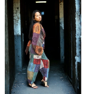 May include: A woman wearing a colorful patchwork pantsuit with a long-sleeved jacket and pants. The fabric is a mix of brown, orange, green, blue, and purple with a geometric pattern. She is standing in a narrow hallway with dark walls.