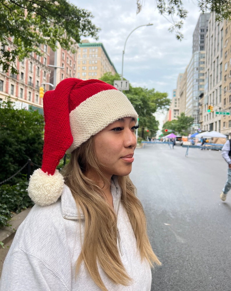 K&ouml;nnte beinhalten: Eine rot-wei&szlig;e gestrickte Weihnachtsm&uuml;tze mit einem wei&szlig;en Bommel oben. Die M&uuml;tze wird von einer Person mit langen blonden Haaren getragen.