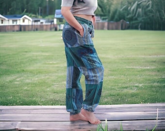 May include: A woman wearing a gray crop top and blue and green patchwork pants with floral designs. She is standing on a wooden platform with green grass in the background.